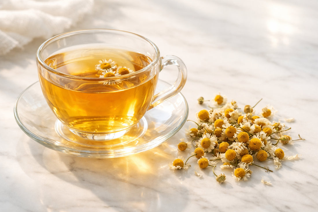 Kamillethee — a clear glass cup of golden chamomile tea with dried chamomile flowers scattered on a white marble surface.